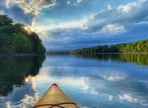 A kayak on calm water reflects a vibrant sky with clouds and trees lining the shore.