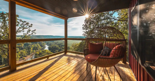 A cozy hanging chair on a wooden deck with a scenic view of hills and a river, illuminated by sunlight.
