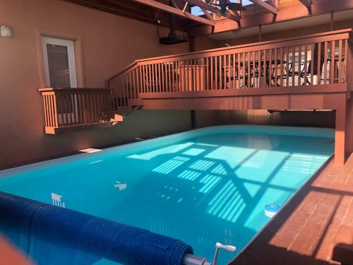 A bright blue swimming pool surrounded by a wooden deck and railing, with sunlight casting shadows on the water.