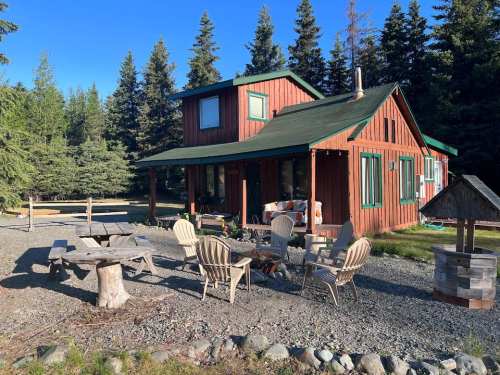 A cozy wooden cabin surrounded by trees, featuring a gravel area with outdoor seating and a fire pit.