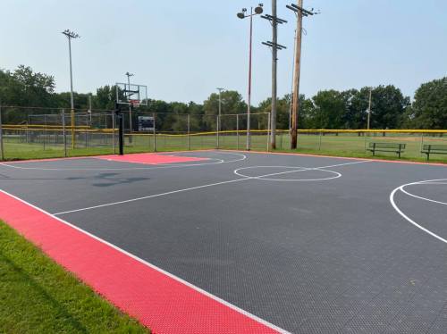 A basketball court with red and gray flooring, surrounded by a grassy area and trees in the background.