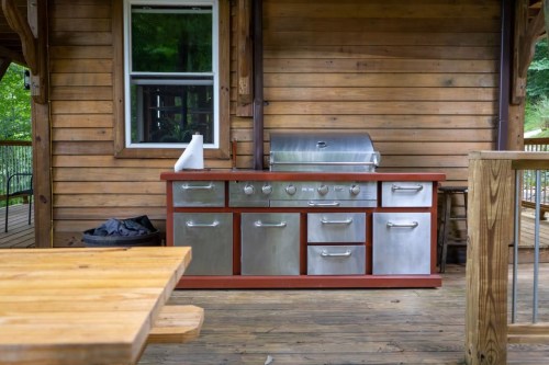 A modern outdoor kitchen with a stainless steel grill and cabinets, set on a wooden deck surrounded by greenery.