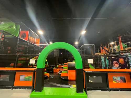 A vibrant indoor trampoline park featuring green and orange structures, with various jumping areas and safety nets.