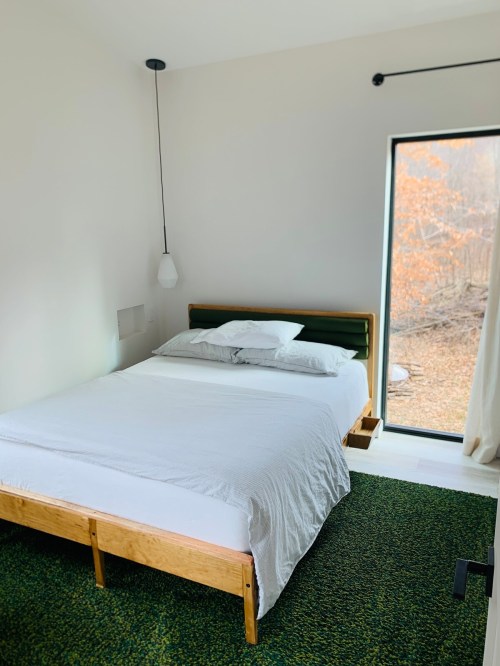 A minimalist bedroom with a wooden bed, white bedding, and a large window overlooking a natural landscape.