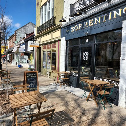 A charming street scene featuring a pasta restaurant with outdoor seating and nearby shops on a sunny day.