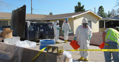 Workers in protective gear handle materials near a dumpster, with caution tape visible in a residential area.