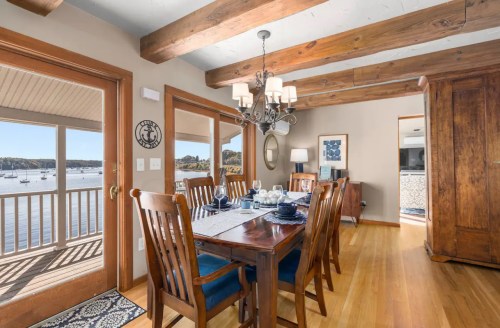 A cozy dining room with wooden beams, a table set for dinner, and a view of the water through large windows.