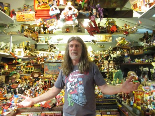 A man with long hair stands in a room filled with colorful toys and collectibles, gesturing with his arms.