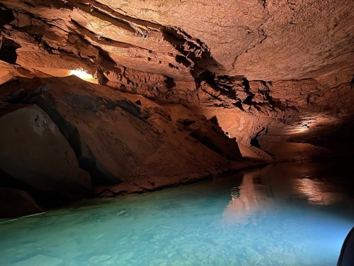 A dimly lit cave with rocky walls and a calm, clear water pool reflecting the surroundings.