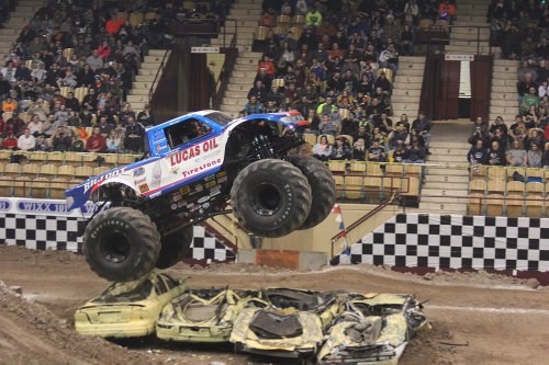 A blue monster truck jumps over a crushed car in a packed arena during a thrilling motorsport event.