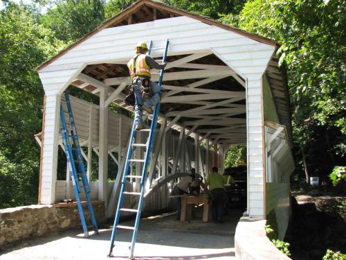 A worker on a ladder repairs a covered bridge, surrounded by trees and construction equipment.