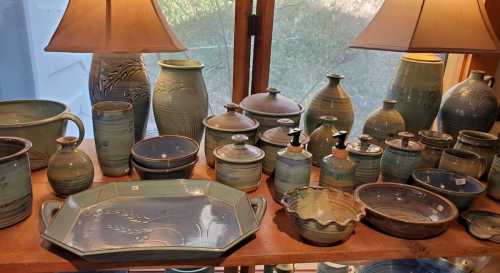 A collection of handcrafted pottery in various shapes and sizes, featuring green and blue glazes, displayed on a wooden shelf.