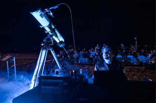 A person operates a telescope at night, surrounded by an audience, with blue lighting and a foggy atmosphere.