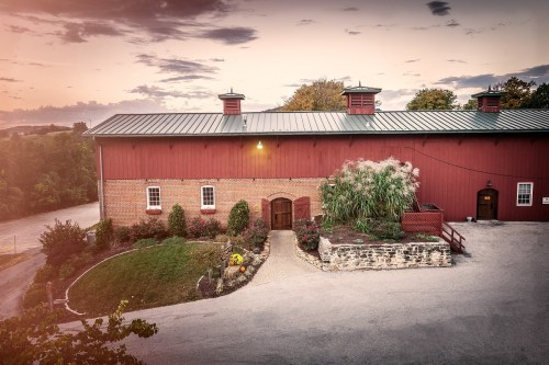 A red barn with a metal roof, surrounded by greenery and a gravel path, under a colorful sunset sky.