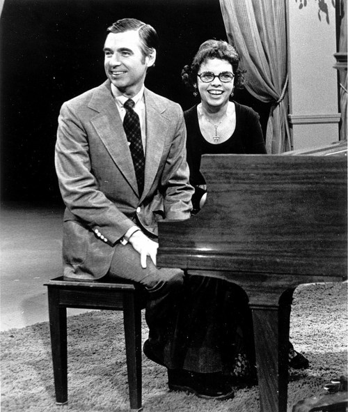 A man and a woman sit at a piano, smiling, in a cozy studio setting with curtains in the background.