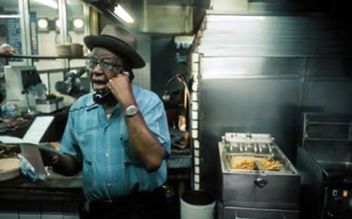 A man in a blue shirt and hat talks on the phone in a busy kitchen, with a fryer and food preparation area visible.