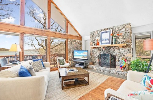 Cozy living room with stone fireplace, large windows, and a view of nature, featuring comfortable seating and a TV.