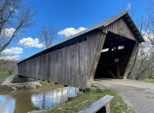 A wooden covered bridge spans a calm stream, surrounded by trees and a blue sky with fluffy clouds.
