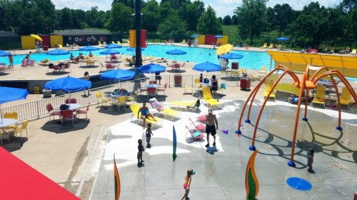 A vibrant outdoor pool area with umbrellas, lounge chairs, and children playing near a water feature on a sunny day.