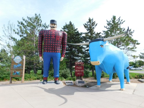 A large statue of Paul Bunyan in plaid, standing next to a blue ox, with trees and a sign in the background.