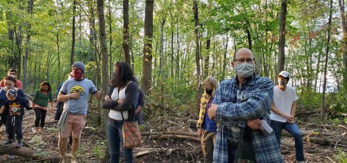 A group of people wearing masks stands in a forest, surrounded by trees, engaged in conversation.