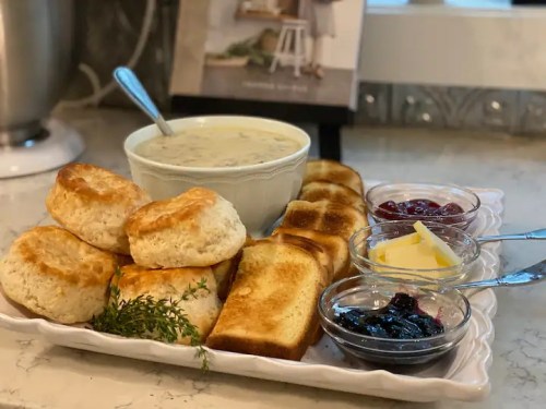 A platter with biscuits, toast, butter, and jams, alongside a bowl of creamy soup, set on a kitchen counter.