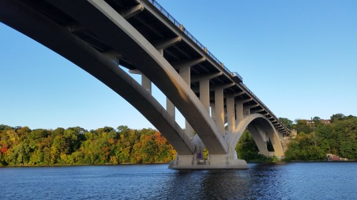 A large concrete bridge arches over a calm river, surrounded by trees with autumn foliage under a clear blue sky.