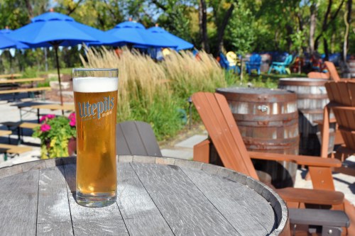 A glass of beer on a wooden table, with outdoor seating and blue umbrellas in the background.