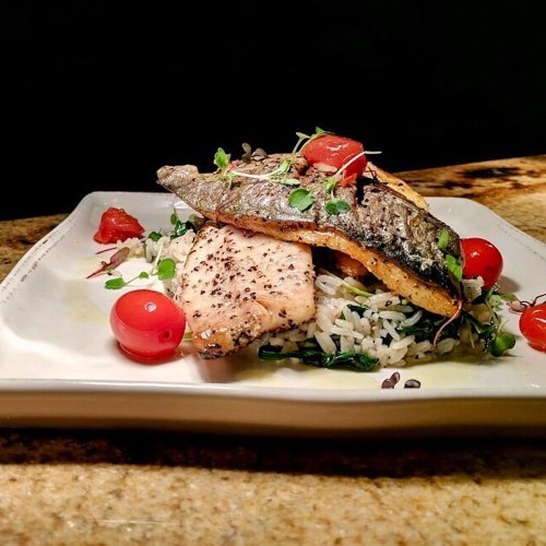 A beautifully plated dish featuring grilled fish, cherry tomatoes, and rice with greens on a white plate.
