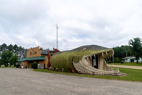 A large, whimsical fish-shaped building serves as a restaurant, set against a cloudy sky and grassy landscape.