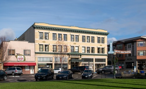 Historic buildings line a street, with parked cars in front and a clear blue sky above.