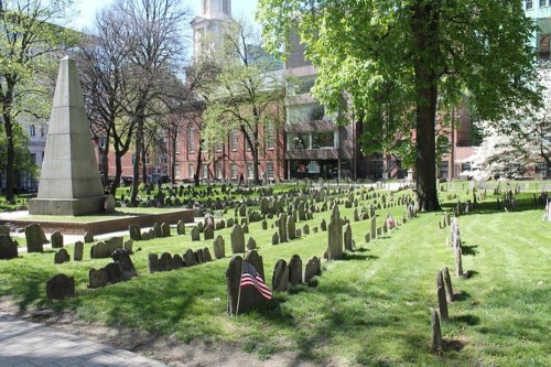 A historic cemetery with numerous gravestones, trees, and a monument, set in a green, peaceful landscape.