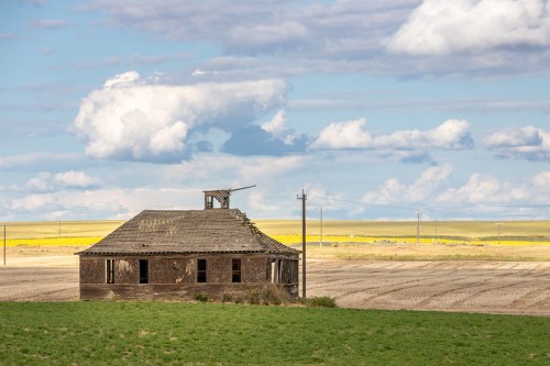 A weathered, abandoned house stands in a vast, open landscape under a blue sky with fluffy clouds.