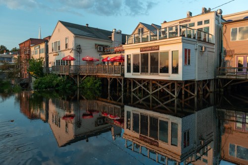 Waterfront buildings with outdoor seating reflect in calm water during sunset.