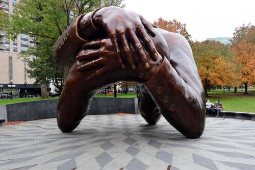 A large bronze sculpture of a pair of hands embracing a torso, set in a park with autumn trees in the background.