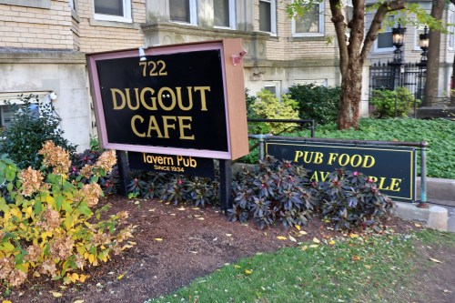 Sign for Dugout Cafe with "Tavern Pub" and "Pub Food Available" surrounded by greenery and flowers.