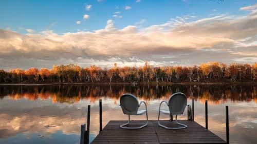 Two chairs on a wooden dock overlooking a calm lake, surrounded by autumn-colored trees and a cloudy sky.