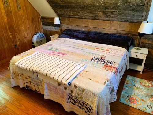 Cozy bedroom with a quilted bedspread, nightstands, lamps, and wooden walls. A fan is visible in the corner.