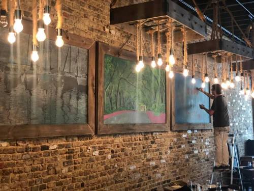 A person on a ladder paints a mural on a brick wall, illuminated by hanging light bulbs.