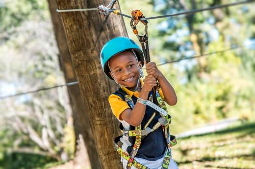 A smiling child in a helmet and harness holds onto a zip line, ready to glide through a forested area.