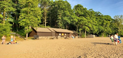 A sandy beach with a wooded area, featuring a building and groups of people enjoying the sun and water activities.