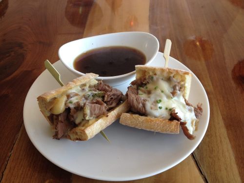 A plate with two beef sandwiches on crusty bread, topped with cheese and served with a small bowl of dipping sauce.