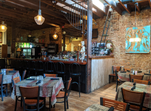 Cozy restaurant interior with wooden beams, a bar, and tables set with patterned tablecloths. Warm lighting creates an inviting atmosphere.