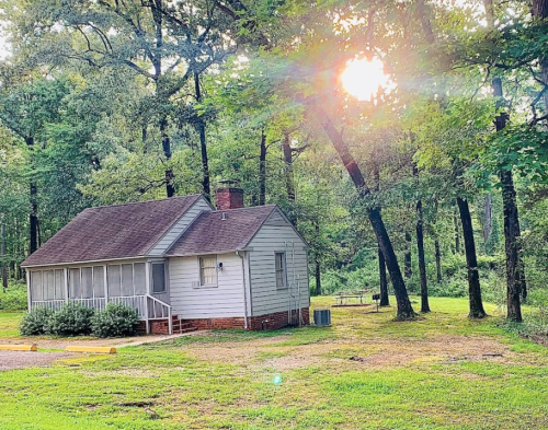 A small, rustic house surrounded by trees, with sunlight filtering through the leaves in a serene outdoor setting.