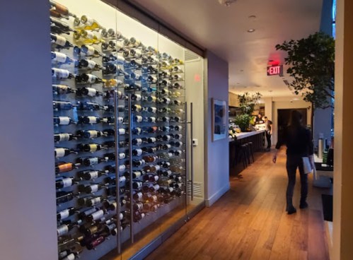 A hallway with a glass wine display on one side and a bar area in the background, featuring wooden flooring.