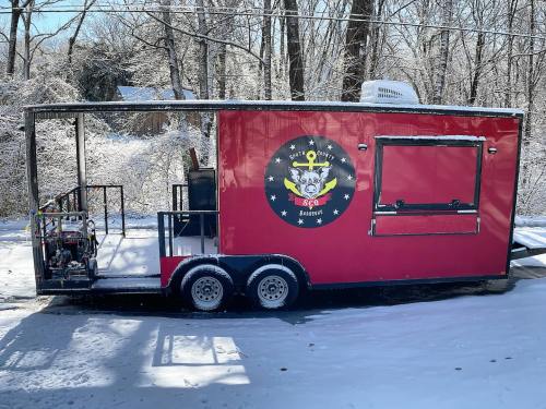 A red food trailer with a logo, parked in a snowy area surrounded by trees.