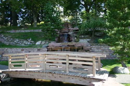 A wooden bridge over a small pond, with a waterfall and lush greenery in the background.
