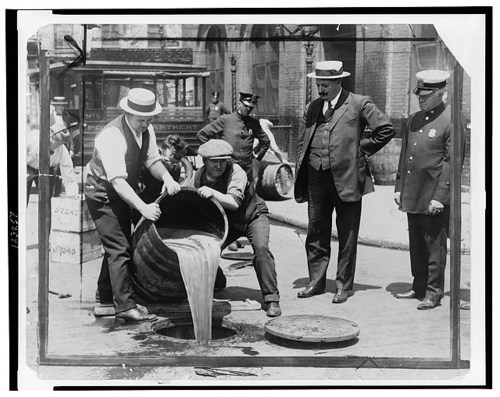 Historical black-and-white photo of men pouring liquid from a barrel into a street drain, with police officers present.