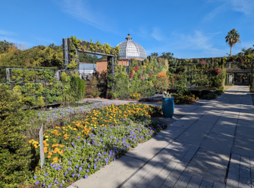 A vibrant garden with colorful flowers, pathways, and a glass dome structure under a clear blue sky.