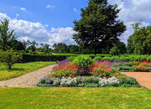 A vibrant garden with colorful flowers, green trees, and a blue sky with fluffy clouds. Paths weave through the landscape.